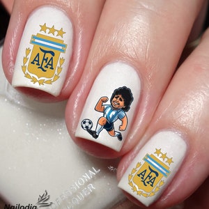 Puede incluir: Esmalte de uñas blanco con arte de uñas azul y dorado con el logotipo de la Asociación Argentina de Fútbol (AFA) y una imagen de dibujos animados de un jugador de fútbol.
