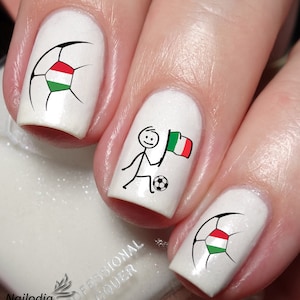 Puede incluir: Esmalte de uñas blanco con arte de uñas de temática futbolística. Una uña tiene un balón de fútbol con la bandera húngara, otra tiene una figura de palo sosteniendo una bandera italiana y un balón de fútbol, y otra tiene un balón de fútbol con la bandera húngara.