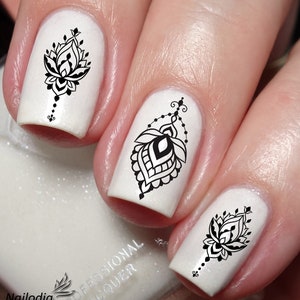 May include: White nail polish with black floral nail art decals. The decals feature intricate designs with a dangling element.