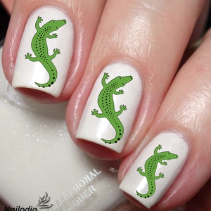 Könnte beinhalten: Drei weiße Fingernägel mit grünen Alligator-Nagelkunst-Aufklebern. Die Alligatoren sind schwarz umrandet und haben schwarze Flecken.