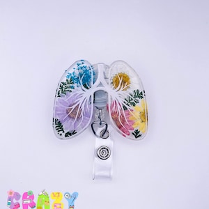 May include: A white resin badge reel with a clear, transparent design featuring a pair of lungs with pressed flowers inside. The flowers are in shades of pink, purple, blue, and yellow.