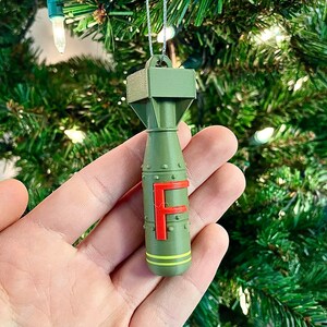 May include: Olive green Christmas ornament shaped like a bomb, with a red "F" on the side. The ornament has yellow stripes and is hanging from a silver string. The background is a Christmas tree.