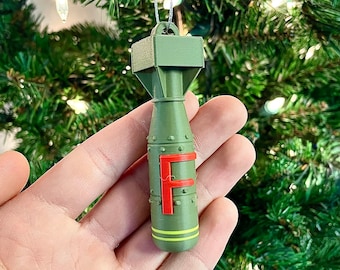 May include: Olive green Christmas ornament shaped like a bomb, with a red "F" on the side. The ornament has yellow stripes and is hanging from a silver string. The background is a Christmas tree.