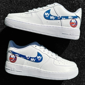 May include: White leather sneakers with a blue and orange New York Islanders logo and a blue and white "NY" swoosh. The sneakers have a white midsole and a white outsole.