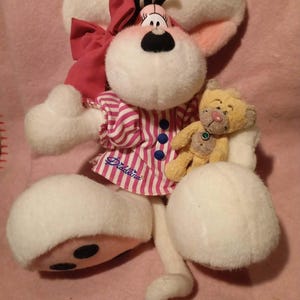 Könnte beinhalten: Ein Diddlina-Maus-Plüschtier mit großen weißen Ohren, einer rosa Schleife und einem rosa-weiß gestreiften Kleid. Die Puppe hält einen kleinen gelben Teddybären. Die Füße der Puppe haben schwarze Punkte.