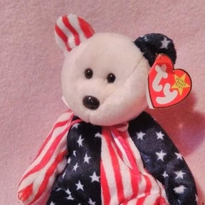 May include: A white Beanie Baby bear wearing a red, white, and blue American flag patterned outfit. The bear has black eyes and a black nose. The bear is sitting on a pink surface. The bear has a Ty tag attached to its ear with the words "Beanie Babies" and a star.