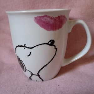 May include: White ceramic mug featuring a cartoon dog design with a kiss mark in pink lipstick. The mug has a curved handle and a simple, clean design. The dog is drawn in black outline.
