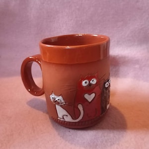 Könnte beinhalten: Eine terrakottafarbene Keramiktasse mit Henkel. Die Tasse hat ein Reliefdesign von drei Katzen in Weiß, Rot und Braun. Der Innenraum und der Rand der Tasse sind in einem dunkleren Terrakottaton gehalten.