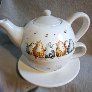 Könnte beinhalten: Ein weißes Keramik-Teeservice für eine Person, bestehend aus einer Teekanne, die auf eine passende Tasse mit Untertasse gestapelt wird. Das Äußere ist mit einer bunten Illustration von fünf verschiedenen Katzen in einer Reihe sowie kleinen Herzen verziert.
