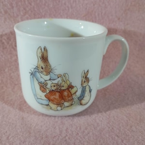 Puede incluir: Taza de cerámica blanca con una encantadora ilustración de conejos al estilo de un cuento. El diseño incluye una madre coneja y sus hijos, con colores suaves y líneas delicadas. La taza tiene un asa curvada y una forma clásica.