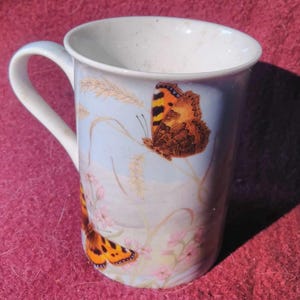 Op de afbeelding: Witte keramische mok met een lichtblauwe achtergrond met twee oranje en zwarte vlinders, tarwearen en roze bloemen. De mok heeft een wit handvat en een witte binnenkant.