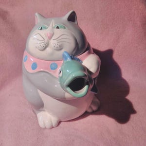 May include: A ceramic teapot shaped like a grey and white cat holding a green fish. The cat has pink and blue polka dots around its neck.