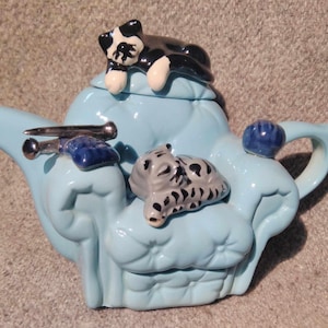 Op de afbeelding: Een lichtblauwe theepot van keramiek in de vorm van een bank met een zwart-witte kat, een grijs-witte kat en een blauwe en zilveren naald op de bovenkant.