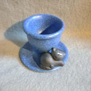 Könnte beinhalten: Ein blau gesprenkelter Eierbecher mit einer kleinen, dunkelgrau geformten Katzenfigur an der Basis. Der Eierbecher hat eine abgerundete Schalenform und einen breiten, kreisförmigen, untertassenartigen Boden, wobei die Katze seitlich sitzt.