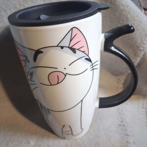Può includere: Una tazza da viaggio in ceramica bianca con un'illustrazione di un gatto dei cartoni animati con orecchie rosa, naso nero e lingua di fuori. La tazza include un coperchio nero e un manico nero. Il design del gatto è realizzato con linee nere.