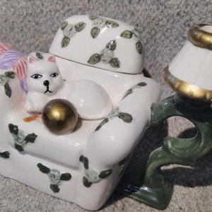 Könnte beinhalten: Weiße Teekanne aus Keramik in Form eines Sessels mit einer Katze, die darin sitzt. Die Teekanne hat ein grün-weißes Blumenmuster und einen goldenen Lampenschirm an der Seite.