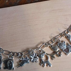 May include: Silver charm bracelet featuring various animal-themed charms, including cats, deer, and bears. The bracelet has a lobster claw clasp and is displayed on a light-colored wooden surface.