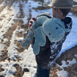 May include: A young person wearing a brown cowboy hat holds a blue crocheted stuffed animal with a textured surface. The person is standing in a snowy outdoor setting.