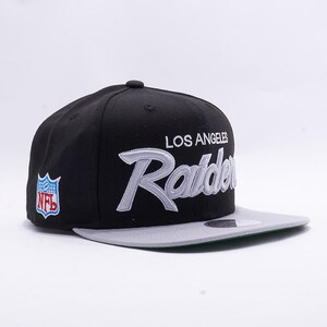 May include: Black and gray snapback hat featuring the Los Angeles Raiders logo in silver script. The hat includes the NFL logo on the side and a gray brim.