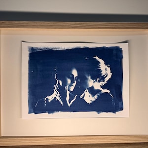 以下が含まれることがあります： 2 人が抱き合っているシアンタイププリントの額縁。画像は薄い青色の色合いで、人物は濃い青色の背景にシルエットで描かれている。