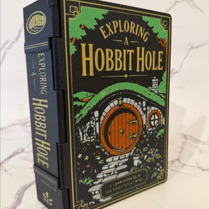May include: A black rectangular deck box with a green and gold illustration of a round door set into a hill. The text reads Exploring A Hobbit Hole and A Commander Deck For Adventurous Minds. The side spine also features the title and a leaf icon.