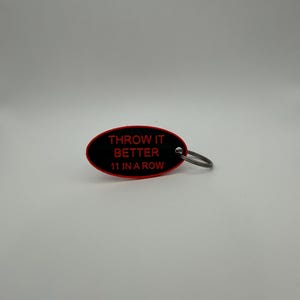 May include: Black and red oval keychain with the text "THROW IT BETTER 11 IN A ROW" in red. The keychain has a silver metal ring attached.