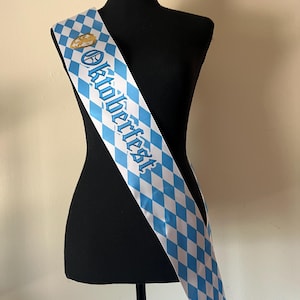 May include: A white and light blue diamond-patterned sash with the word "Oktoberfest" in blue script. A gold-colored emblem is near the top. The sash is draped over a black mannequin.
