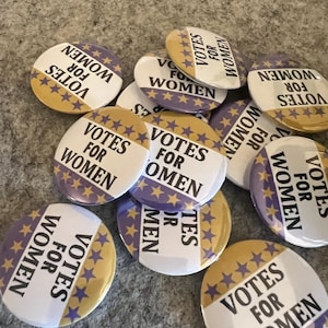 May include: A collection of round buttons with the text "VOTES FOR WOMEN" in white on a purple and gold background. The buttons feature a star design and are scattered across a textured surface.