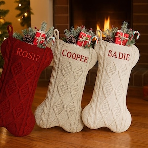 May include: Three Christmas stockings in red, beige, and cream, each with a cable knit design and the names "Rosie", "Cooper", and "Sadie" embroidered on them. Stockings are filled with festive decorations.