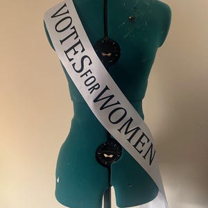 May include: A white sash with the text "VOTES FOR WOMEN" in black lettering is draped over a teal dress form. The sash is angled across the form, with the text clearly visible. The dress form is on a stand.