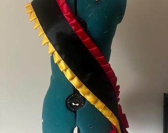 Op de afbeelding: Een zwarte sjerp met rode en gele randen, gedrapeerd over een turquoise paspop. De sjerp heeft een centrale zwarte band met rode en gele gefranjerde randen. De kleuren zijn levendig en de sjerp lijkt van satijn te zijn.