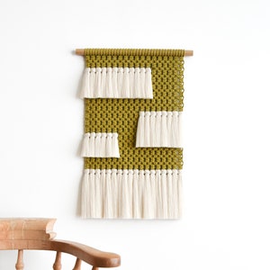 May include: A wall hanging made of woven macrame with a geometric pattern in mustard yellow and white. The hanging has a fringe of white yarn.