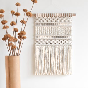 May include: A white macrame wall hanging with a geometric pattern and fringe. The hanging is attached to a wooden dowel rod.