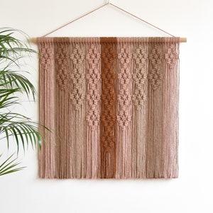 May include: A macrame wall hanging featuring a geometric design in shades of brown and pink. The hanging is crafted from natural fibres and has a rustic, bohemian aesthetic.