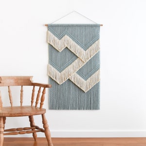 Puede incluir: Un tapiz de macramé de pared con un patrón geométrico de hilo azul y blanco. El tapiz está hecho con una serie de nudos y flecos.