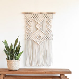 May include: A white macrame wall hanging with a geometric pattern and fringe. The hanging is mounted on a wooden dowel and is displayed on a light brown wooden bench.