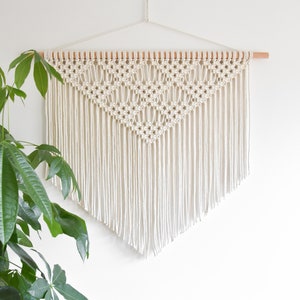 Op de afbeelding: Een witte macramé wandhanger met een geometrisch patroon en franjes. De hanger is gemaakt van natuurlijke katoenen koord en is bevestigd aan een houten lat.