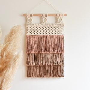 Könnte beinhalten: Ein Makramee-Wandbehang mit geometrischem Muster und einem mehrstufigen Fransen in Beige- und Rosatönen.