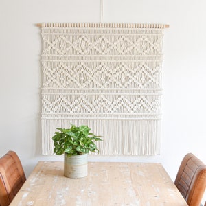 May include: A white macrame wall hanging with a geometric pattern and fringe. The hanging is mounted on a wooden dowel rod and is displayed on a white wall above a wooden table with a potted plant.