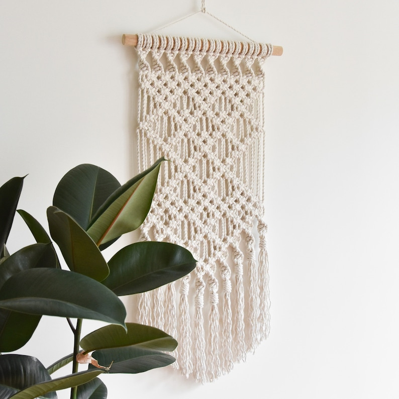 May include: A white macrame wall hanging with a diamond pattern and fringe. The hanging is made of cotton cord and is attached to a wooden dowel.
