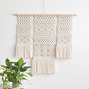 Peut inclure: Une suspension murale en macramé blanche avec trois panneaux, chacun avec un motif géométrique différent. Les panneaux sont reliés par une baguette en bois.