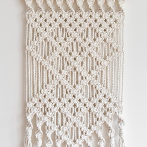 May include: A white macrame wall hanging with a diamond pattern and fringe tassels.
