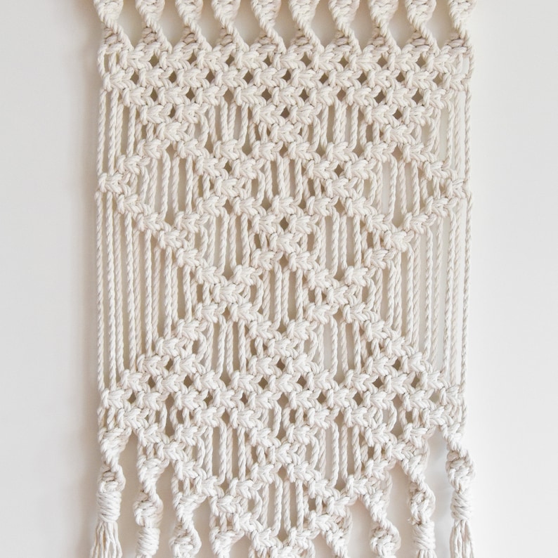 May include: A white macrame wall hanging with a diamond pattern and fringe tassels.