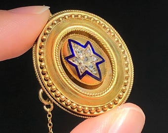 May include: A round gold brooch featuring a central six-pointed star inlay with blue enamel borders and small clear stones. The brooch has a beaded gold edge and a delicate gold chain attached to the bottom, set against a black background.