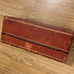 K&ouml;nnte beinhalten: Eine rote Lederbox mit goldenen Verzierungen und einem gewellten Rand. Die Box ist geschlossen und hat einen Klappdeckel.