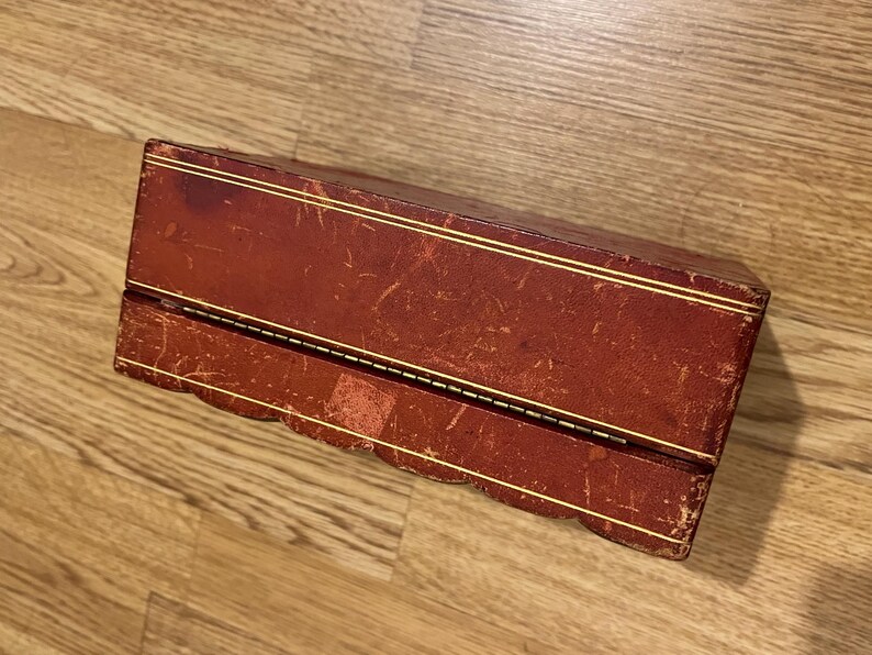 K&ouml;nnte beinhalten: Eine rote Lederbox mit goldenen Verzierungen und einem gewellten Rand. Die Box ist geschlossen und hat einen Klappdeckel.