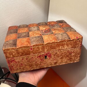 K&ouml;nnte beinhalten: Eine antike Holzkiste mit einem Schachbrettmuster auf dem Deckel. Die Kiste ist aus braunem und orangefarbenem Holz und hat ein Schl&uuml;sselloch an der Vorderseite. Die Kiste ist leicht abgenutzt und hat einen Used-Look.