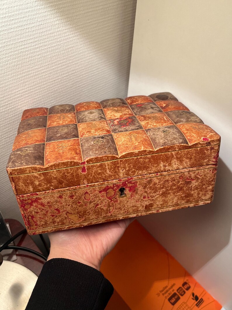 K&ouml;nnte beinhalten: Eine antike Holzkiste mit einem Schachbrettmuster auf dem Deckel. Die Kiste ist aus braunem und orangefarbenem Holz und hat ein Schl&uuml;sselloch an der Vorderseite. Die Kiste ist leicht abgenutzt und hat einen Used-Look.