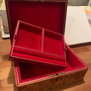 K&ouml;nnte beinhalten: Eine Schmuckschatulle aus rotem Samt mit goldener Verzierung und einem gemusterten &Auml;u&szlig;eren. Die Schachtel hat zwei F&auml;cher im Inneren, die mit rotem Samt ausgekleidet sind.