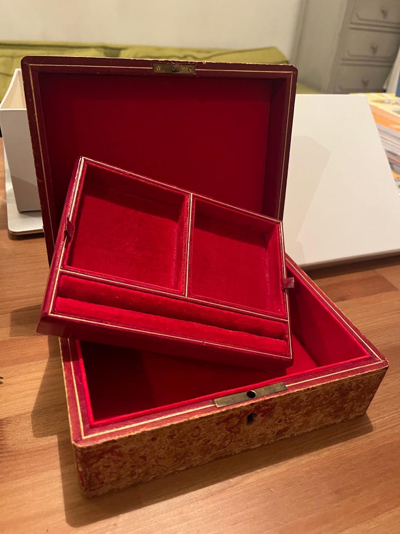 K&ouml;nnte beinhalten: Eine Schmuckschatulle aus rotem Samt mit goldener Verzierung und einem gemusterten &Auml;u&szlig;eren. Die Schachtel hat zwei F&auml;cher im Inneren, die mit rotem Samt ausgekleidet sind.
